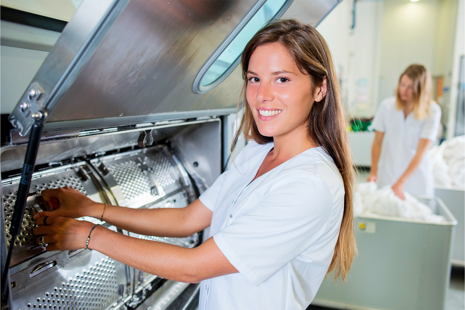 Lavoratrice di una lavanderia industriale sorride verso la camera mentre sta chiudendo lo sportello di una lavatrice industriale