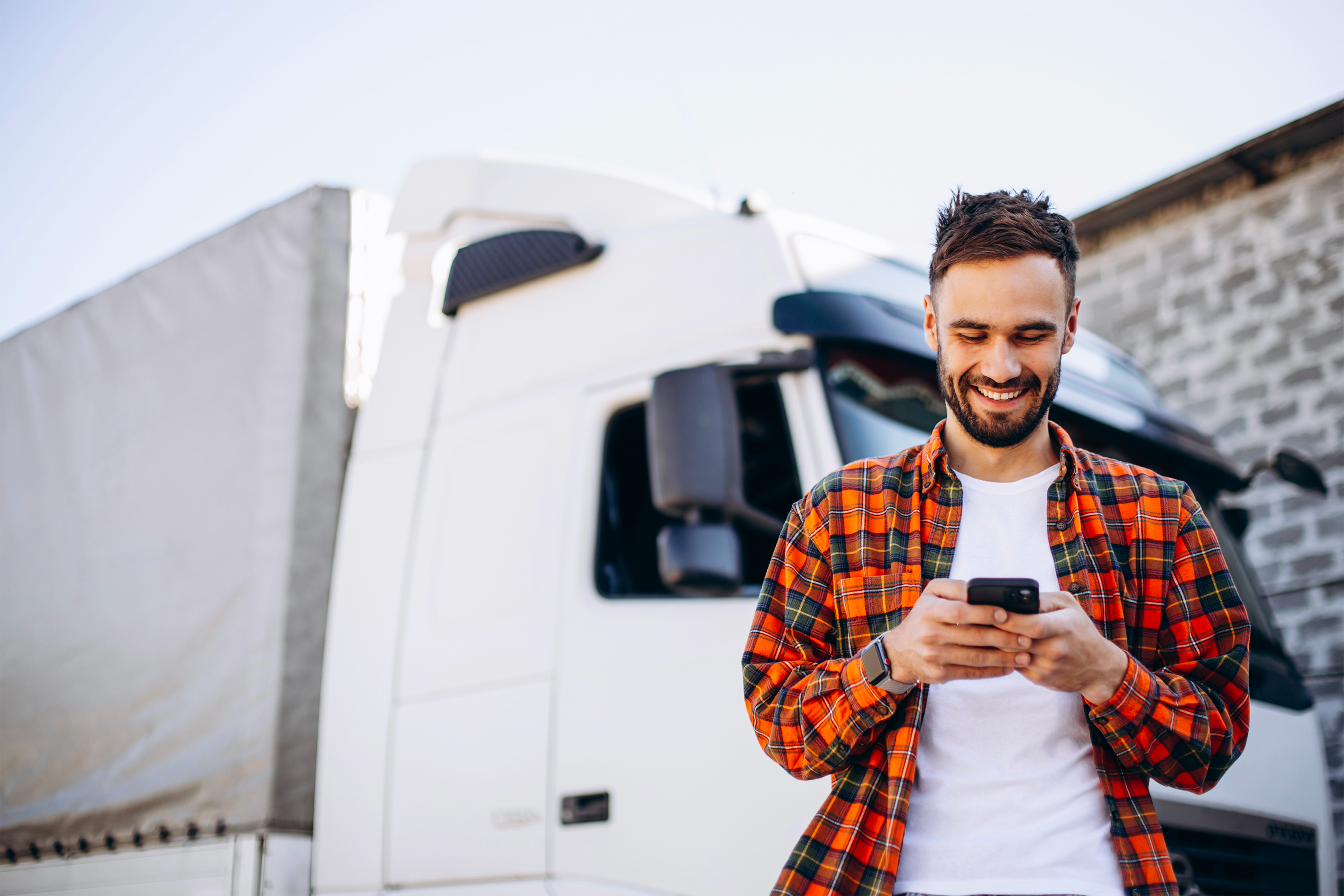 Autista di Camion sorride mentre usa app su smartphone. Si trova all'aperto, con luce diurna con un camion alle sue spalle.