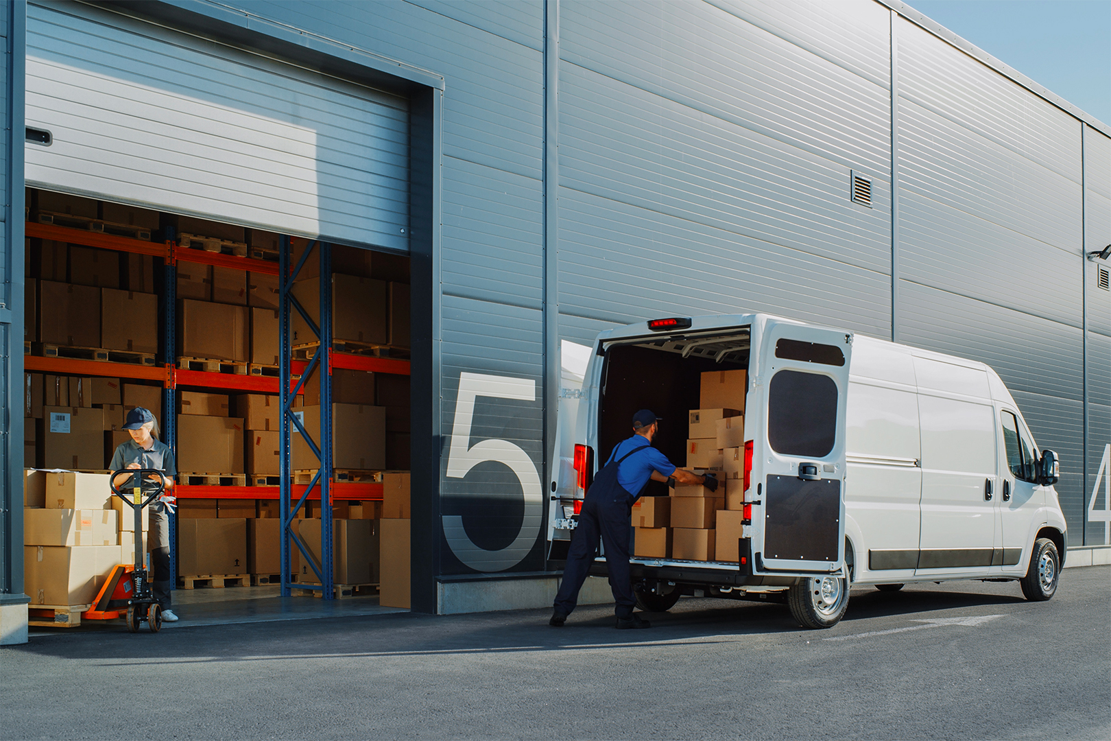 Immagine di un magazzino di logistica ripreso dall'esterno dove due addetti stanno caricando pacchi dentro un furgone.