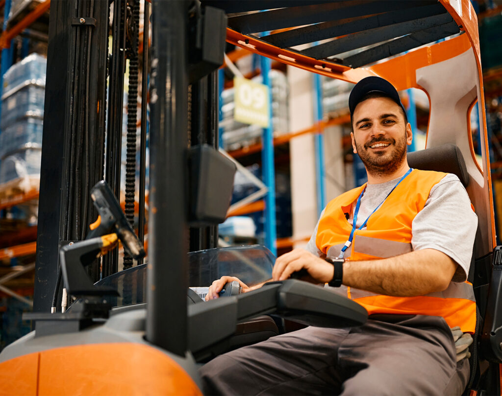 Autista di muletto sorride a bordo di un muletto. La scena si svolge all'interno di un magazzino.