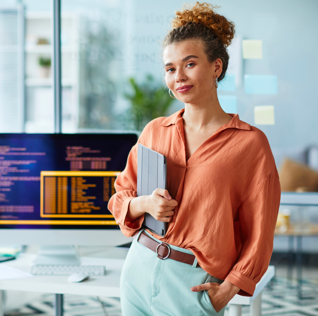 Una programmatrice guarda verso l'obbiettivo. Ha in mano un tablet chiuso. Sullo sfondo un computer con del codice in un luminoso ambiente open space.