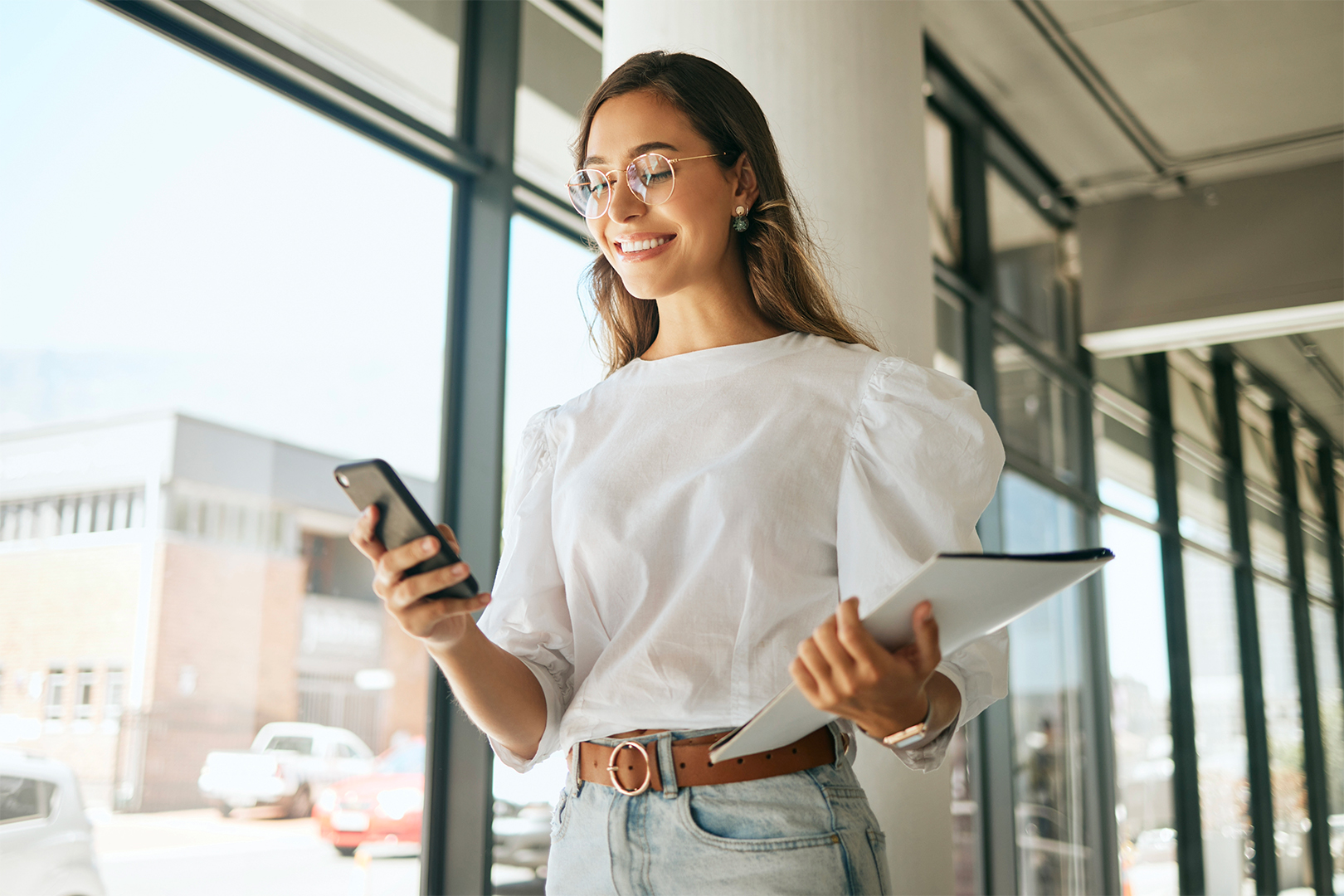 Giovane prosessionista, controlla lo smartphone mentre cammina sorridendo nel corridoio della sede di un'azienda.
