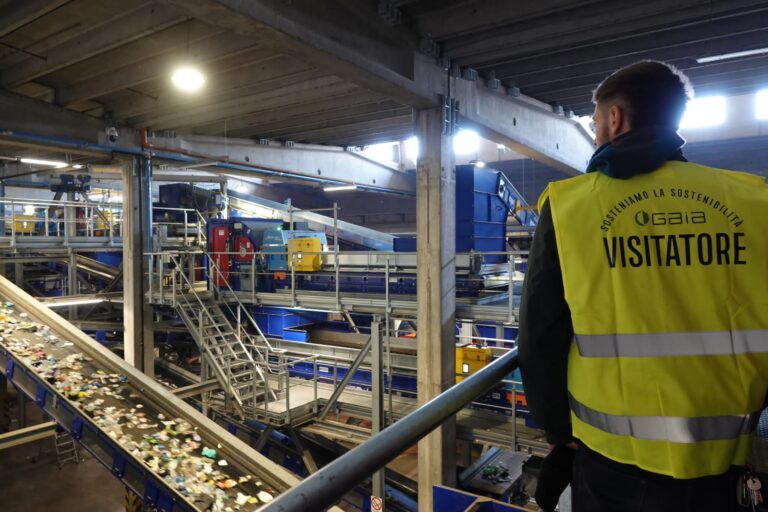 Veduta dall'alto dell'interno di un impianto di trattamento rifiuti con nastri trasportatori carichi di materiali plastici e cabine di selezione automatizzata. In primo piano, un visitatore con gilet giallo osserva il processo.
