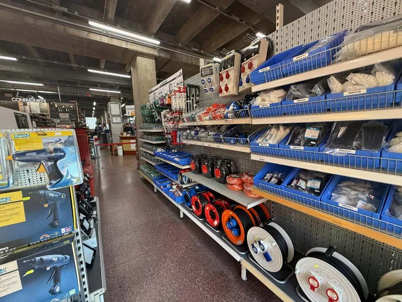 Scaffalature industriali organizzate con vaschette blu contenenti viteria, utensili elettrici in esposizione e rotoli di cavi ordinati in un magazzino commerciale.