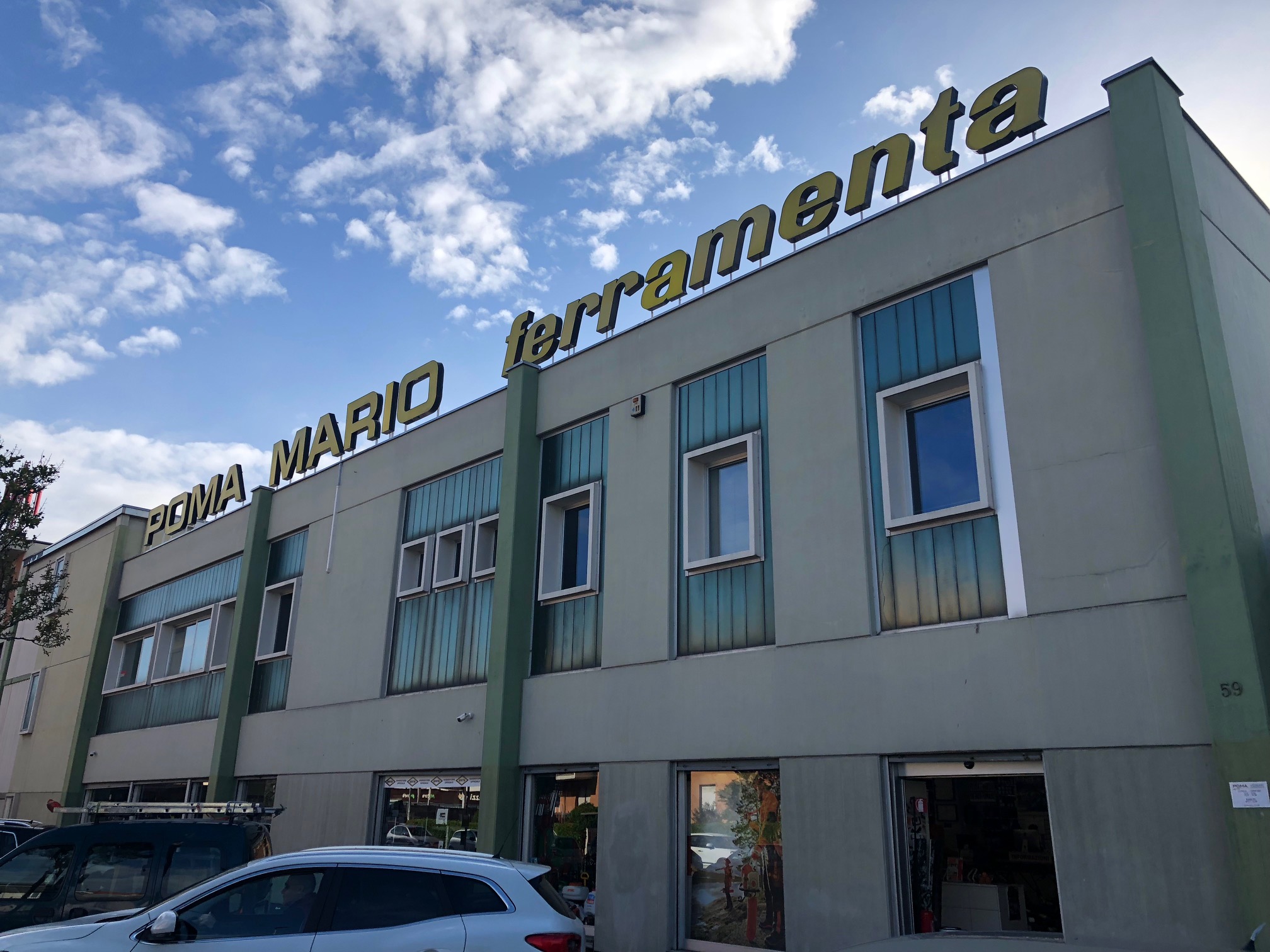 Facciata esterna dell'edificio commerciale Poma Mario Srl con grande insegna a lettere gialle e ampie vetrate, sotto un cielo parzialmente nuvoloso.